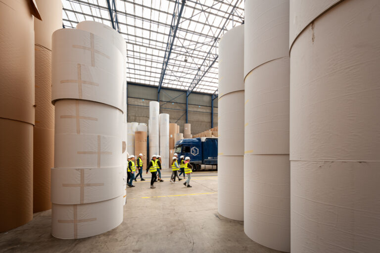 Groep cursisten volwassenenonderwijs wandelen door een magazijn met rollen verpakt materiaal tijdens een bedrijfsbezoek in de haven. © Kris Jacobs