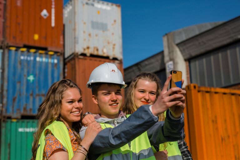 Drie studenten hoger onderwijs in veiligheidshesje, waarvan één met een helm, maken samen een selfie voor gestapelde containers tijdens hun haventour. © Jesse Willems
