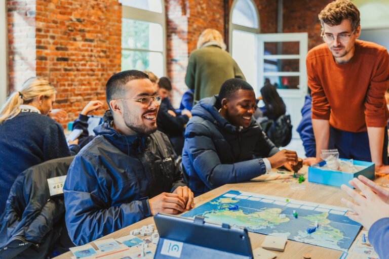 Twee leerlingen secundair onderwijs zitten aan tafel en spelen het Port Xpert Shipping-spel. Ze krijgen begeleiding van een educoach en glimlachen. © Havenwereld